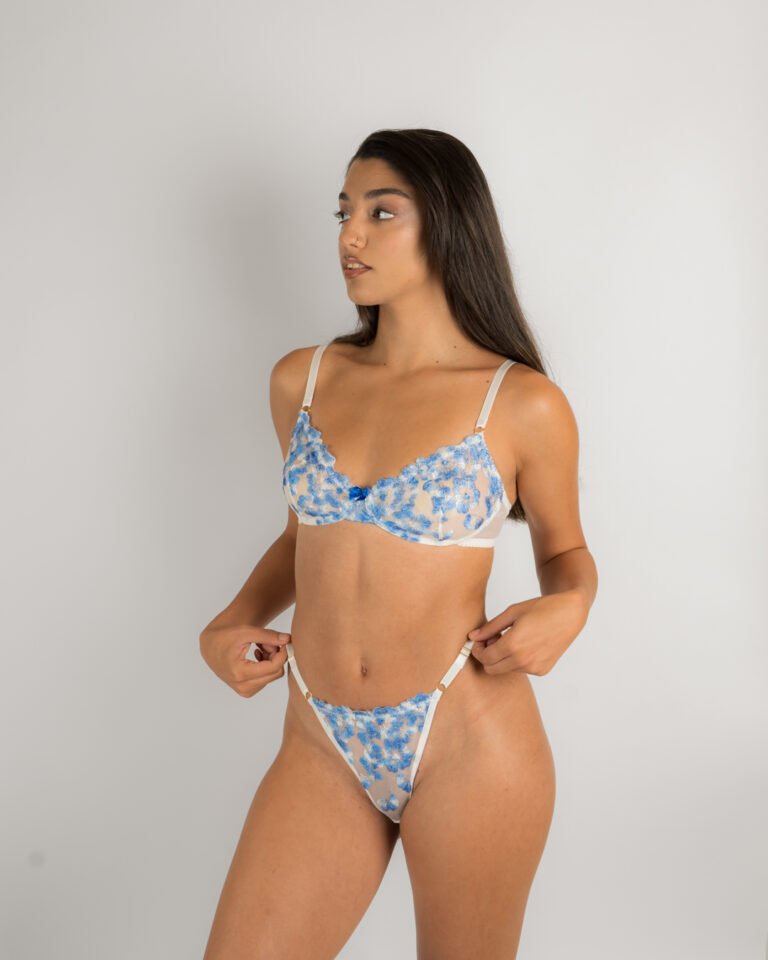 סט הלבשה תחתונה של מותג ההלבשה התחתונה של דנה טנא, הסט בצבע לבן עם רקמת פרחים כחולים ופפיון מתוק. החזיה רכה ונוחה והתחתון בגזרת חוטיני סקסי. תמונת עמידה קדמית